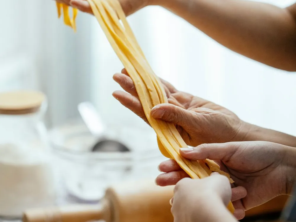 Hands of different generations passing freshly cut tagliatelle to each other, with the rolling pin and the sfoglia on the board