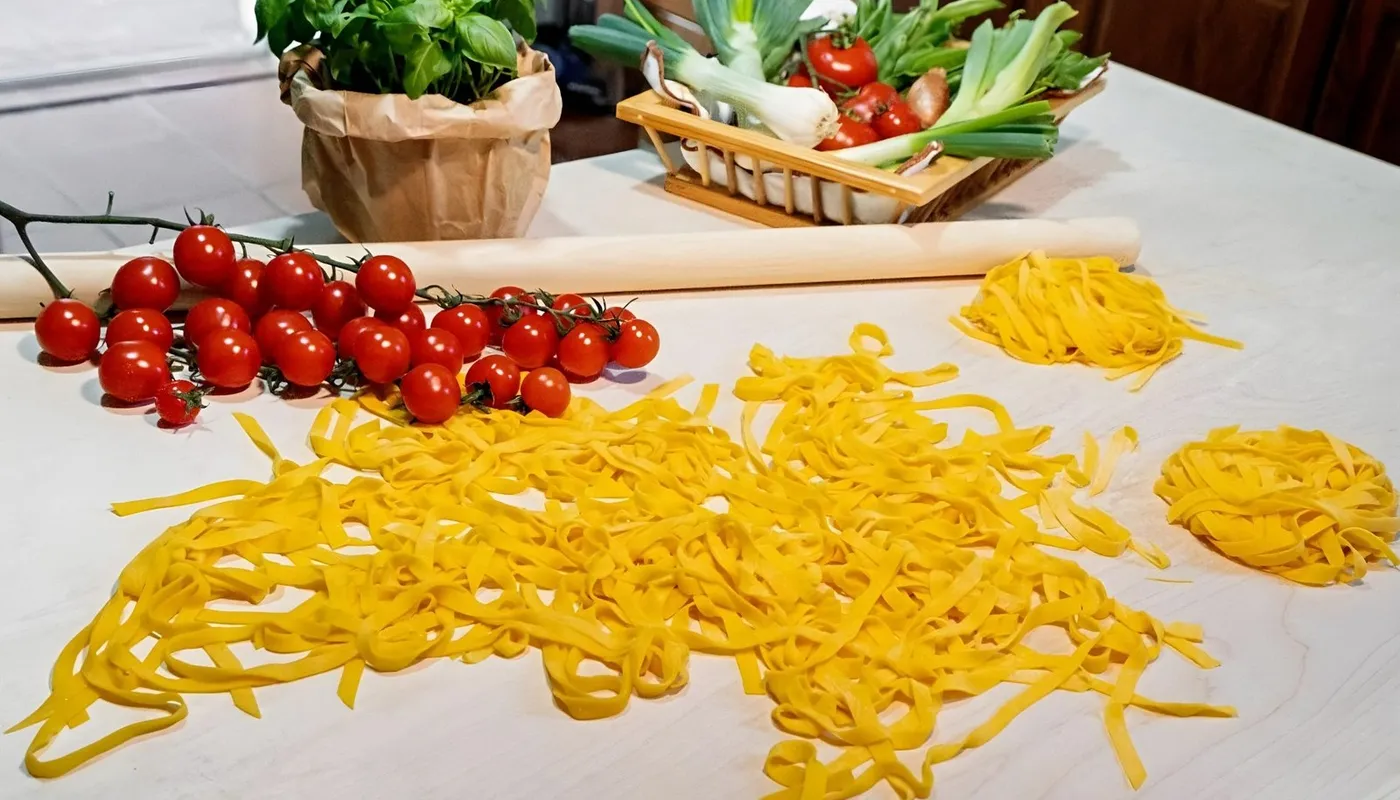 Tagliatelle appena tagliate sparse sul tavolo, con i nidi, il mattarello, pomodorini e basilico