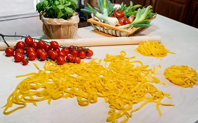 Tagliatelle appena tagliate sparse sul tavolo, con i nidi, il mattarello, pomodorini e basilico