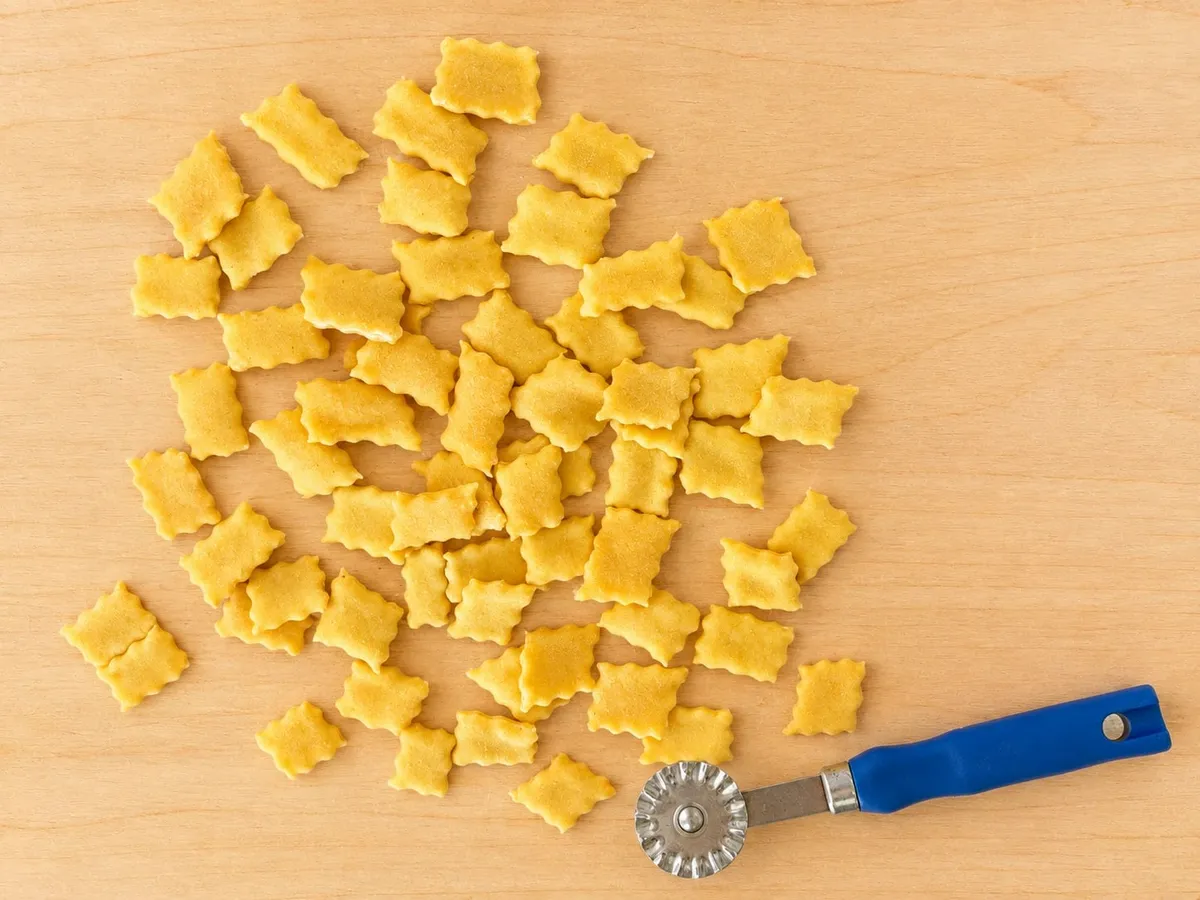 Little squares of spoja lorda with a scalloped edge scattered on the board, next to the fluted pastry wheel
