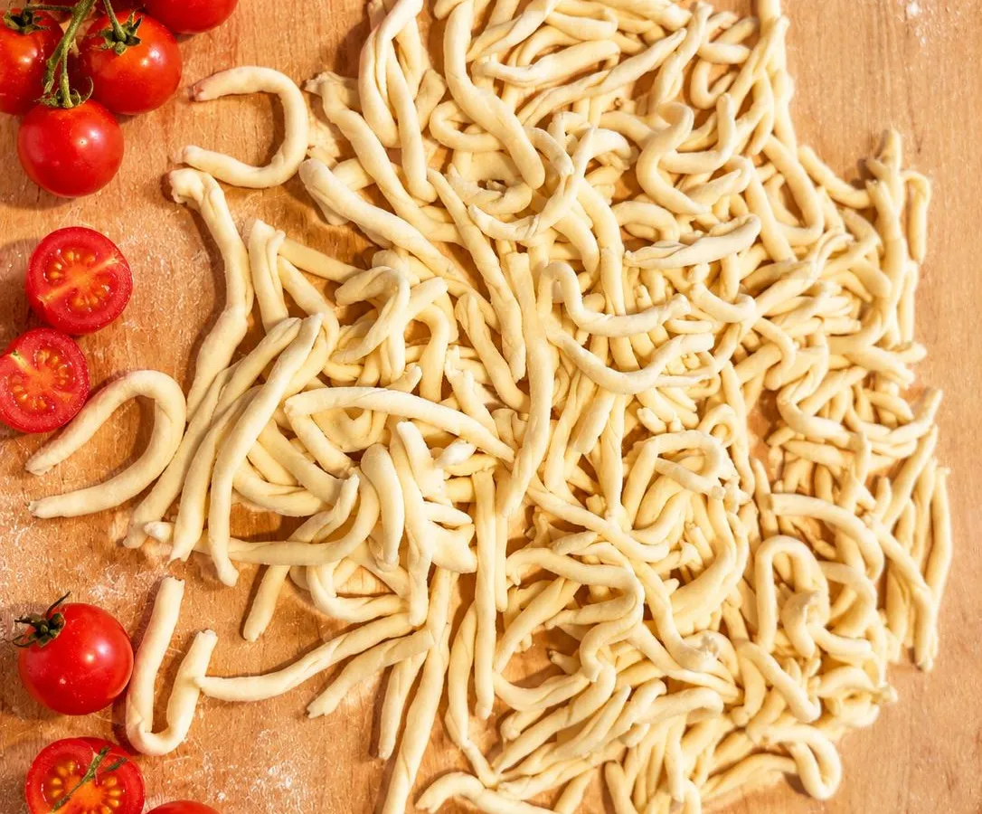 Strozzapreti crudi arrotolati a mano su un tagliere di legno infarinato, con pomodorini a fianco