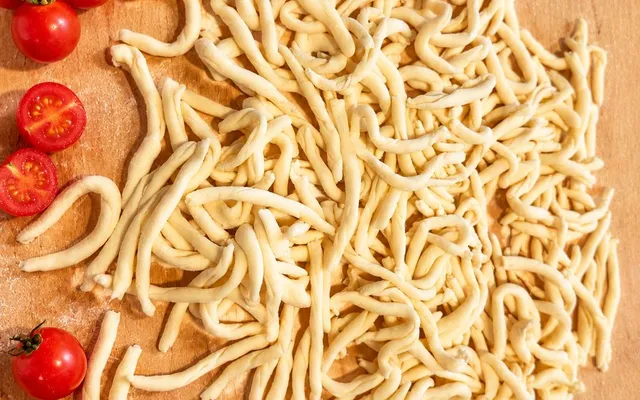 Strozzapreti crudi arrotolati a mano su un tagliere di legno infarinato, con pomodorini a fianco