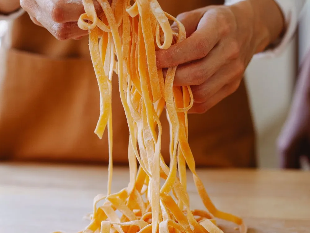 Mani infarinate che sollevano dal tagliere un mazzo di tagliatelle appena tagliate, davanti a un grembiule arancione